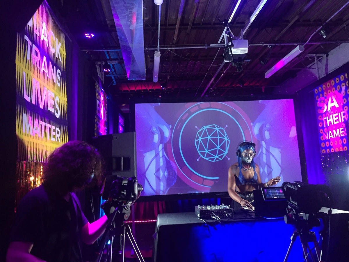 Kha'DJ, wearing long blue wig, head phones, and a mask, stands at a turntable playing music. Projector screens surround the DJ, displaying blue and purple lights and the sayings Black Trans Lives Matter and Say Their Names. To the left is a camera man working a camera that is recording the DJ.