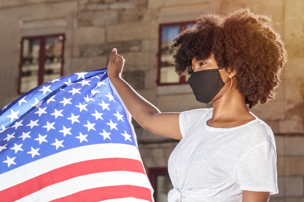 A woman with brown skin wears a black mask and holds an American flag.