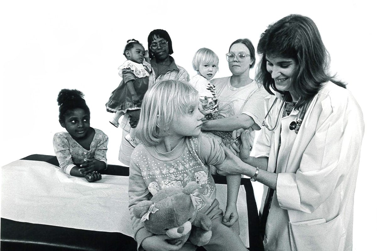 Black and white photo of doctor giving a shot to a child with families in the background.