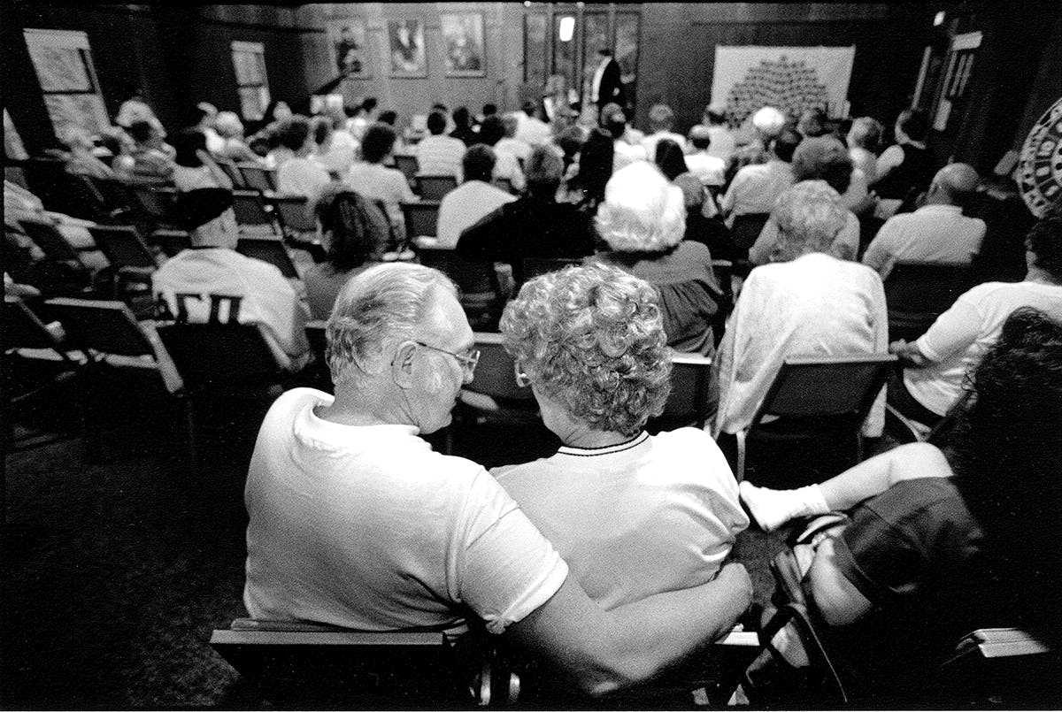 Black and white photo of elderly couple in foreground of a full room at a community meeting.