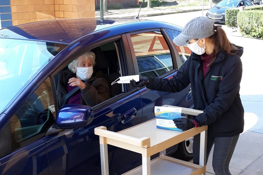 A masked woman hands a mask to another masked woman through a driver side window.